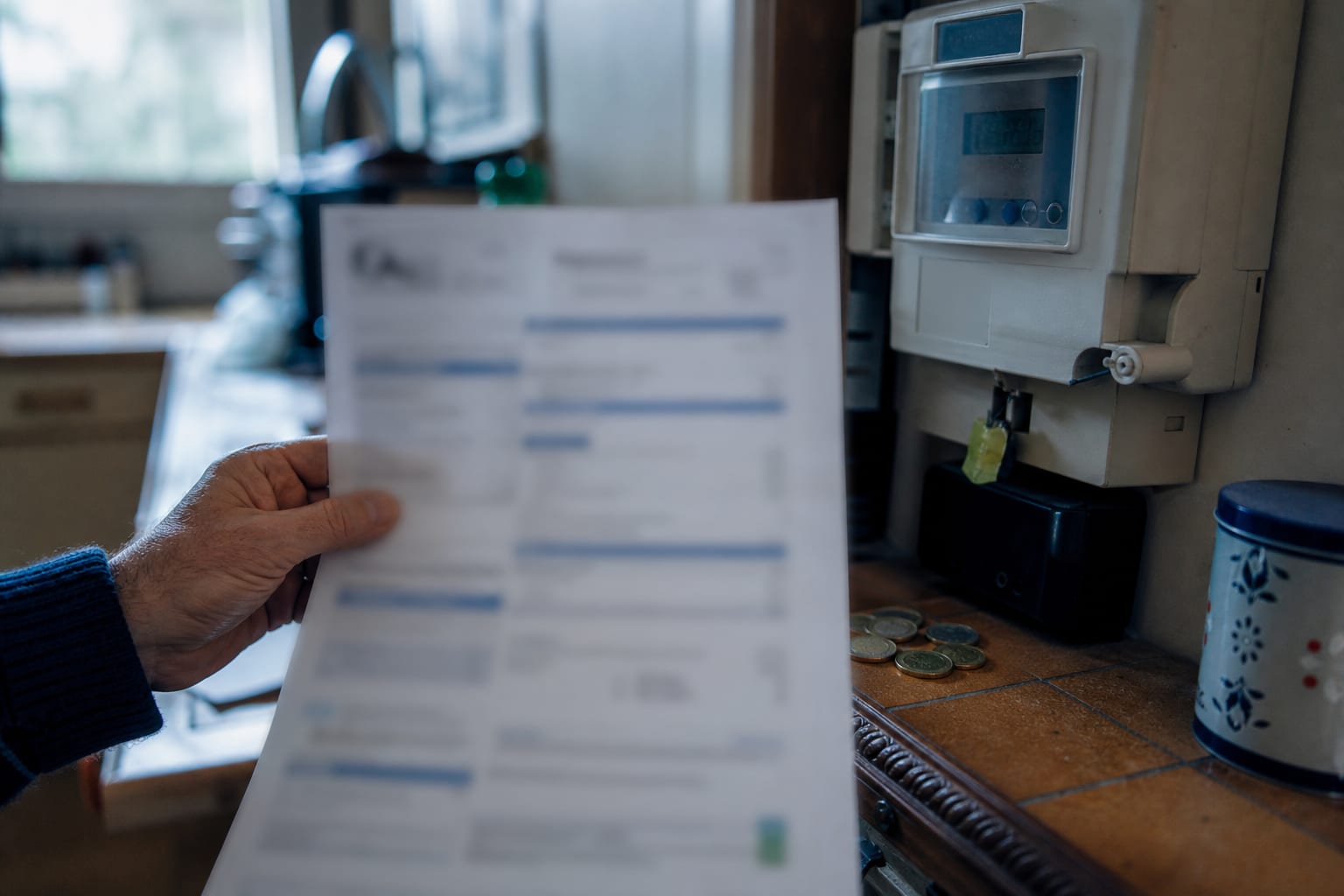 Main tenant une facture floue près du compteur et de quelques pièces.